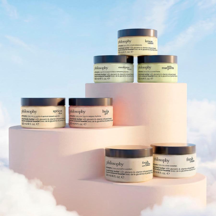 Six Philosophy haircare jars arranged on pastel pink display blocks against a soft blue sky background with clouds. The jars have dark lids and cream labels with different product names including neoyplica, buba, and margita.