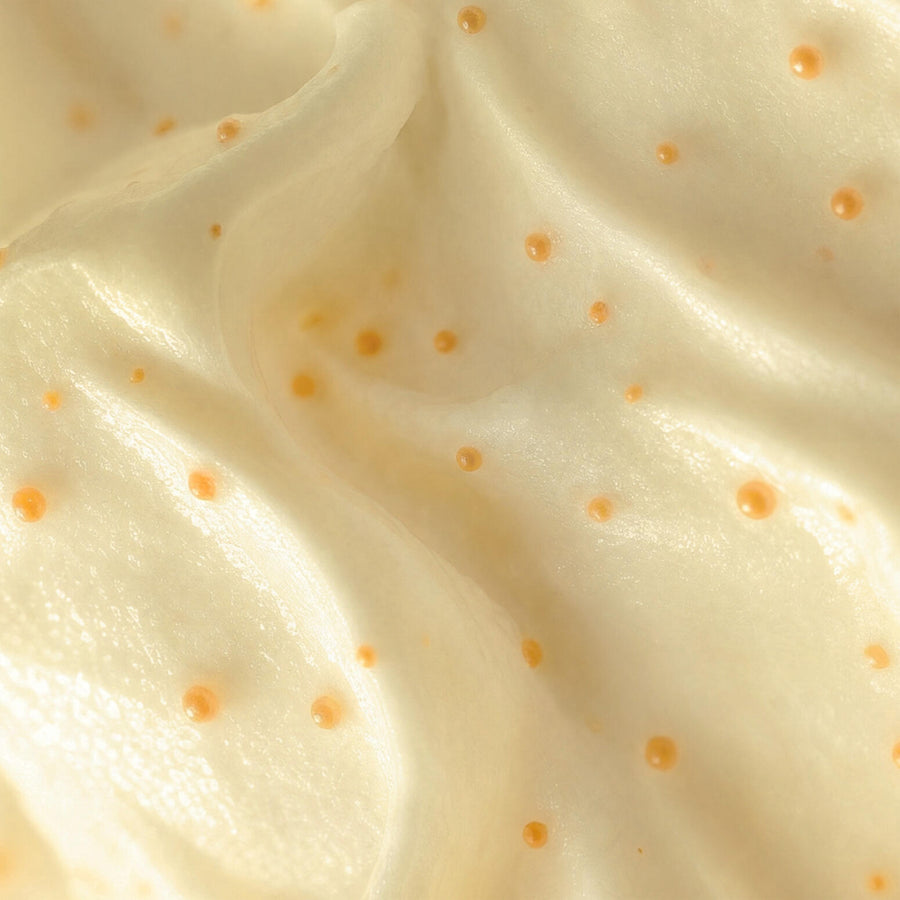 A close-up texture shot of a creamy, off-white whipped product, likely a cosmetic cream or whipped lotion, swirled with small, scattered amber-orange droplets.