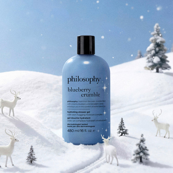 A bottle of Philosophy Blueberry Crumble Hydrating Shower Gel stands in a snowy winter landscape with white deer and small evergreen trees. The bottle is light blue with a black cap, and the background shows snow-covered hills under a pale blue sky.