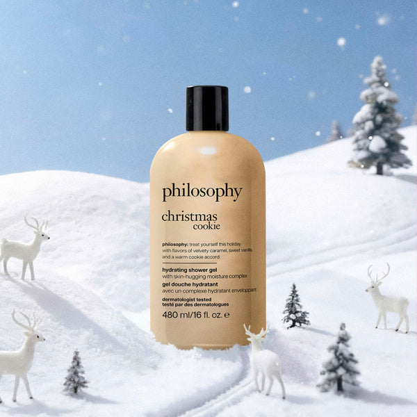 A bottle of Philosophy Christmas Cookie hydrating shower gel stands in a snowy winter scene with white deer and small pine trees. The bottle is a warm beige/tan color with a black cap, and the background is a blue-toned snowy landscape.