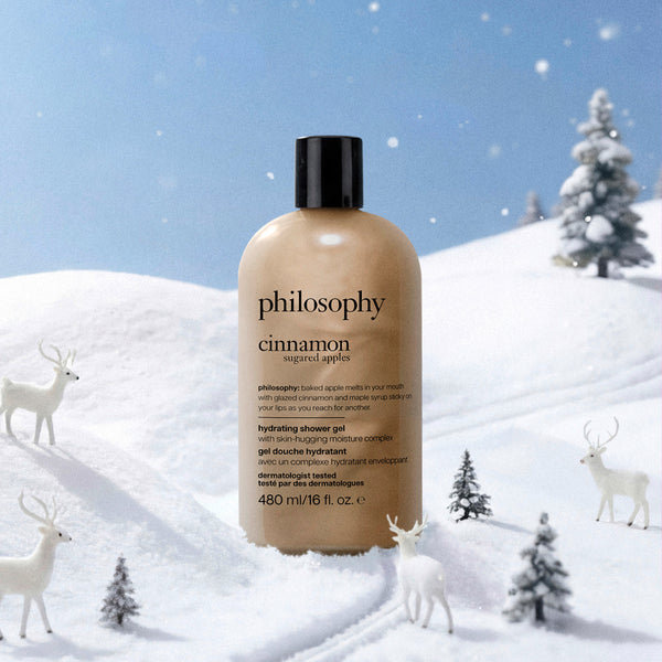 A bottle of Philosophy Cinnamon Sugared Apples hydrating shower gel stands in a snowy winter landscape with deer and small trees. The bottle is a warm beige/cream color with a black cap, and the background shows a blue sky and white snow.
