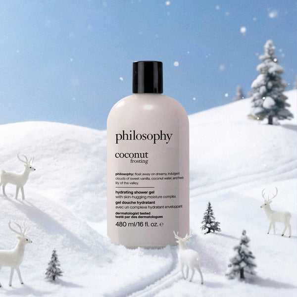 A bottle of Philosophy Coconut Frosting hydrating shower gel stands in a snowy winter scene with white deer and small pine trees. The bottle is off-white with a black cap, and the background shows a blue sky and snow-covered hills.