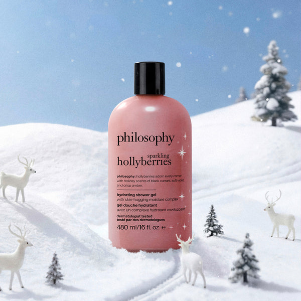 A pink bottle of Philosophy Hollyberries Hydrating Shower Gel stands in a snowy winter scene with white deer and small pine trees. The bottle has a black cap and black text, and the background shows a blue sky and snow-covered ground.