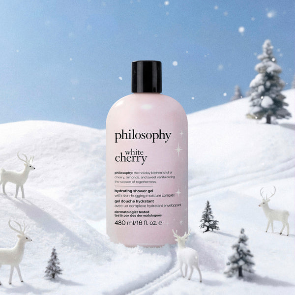 A bottle of Philosophy White Cherry Hydrating Shower Gel stands in a snowy winter landscape with deer and small trees. The bottle is pale pink with a black cap, and the background shows a blue sky and snow-covered hills.
