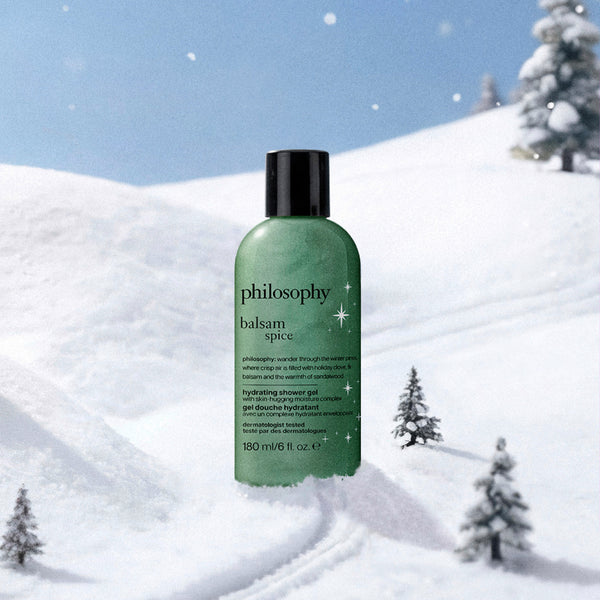 A green bottle of Philosophy Balsam Spice hair balm with a black cap, set against a snowy winter landscape with pine trees under a blue sky.