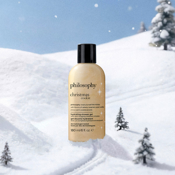 A bottle of Philosophy Christmas Cookie dehydrating shower gel stands against a snowy winter landscape with snow-covered hills, evergreen trees, and a blue sky.