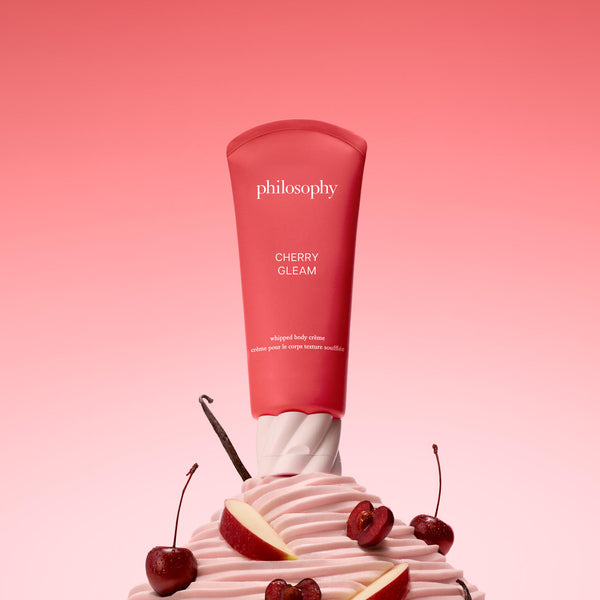 Philosophy Cherry Glem whipped body cream in a pink squeeze tube, styled on top of a pink whipped cream swirl with cherry slices and cherics against a pink gradient background.