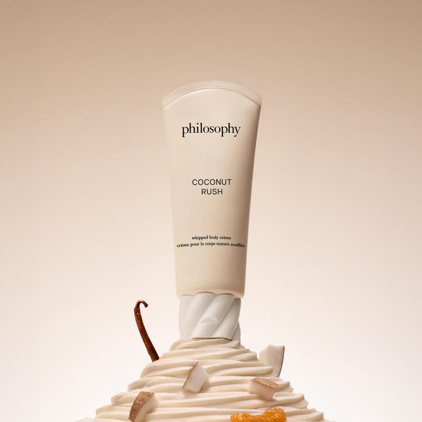 Philosophy Coconut Rush whipped body cream in a white squeeze tube, displayed upright on a sculpted swirl of cream with small coconut and orange pieces against a warm beige background.