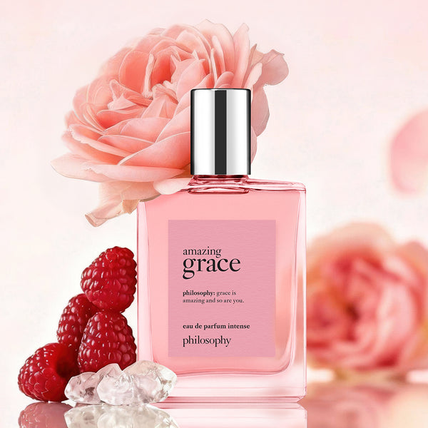 A bottle of Philosophy 'Amazing Grace' Eau de Parfum Intense, filled with pink liquid and featuring a silver cap, styled with a large pink rose, fresh raspberries, and clear crystals on a reflective surface against a soft pink background.