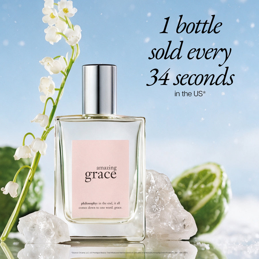 A clear rectangular perfume bottle labeled 'amazing grace' with a pale pink front label and a silver cap, styled on a reflective surface with white flowers, musk crystals, and green citrus against a light blue background. Text on the image reads '1 bottle sold every 34 seconds in the US'.