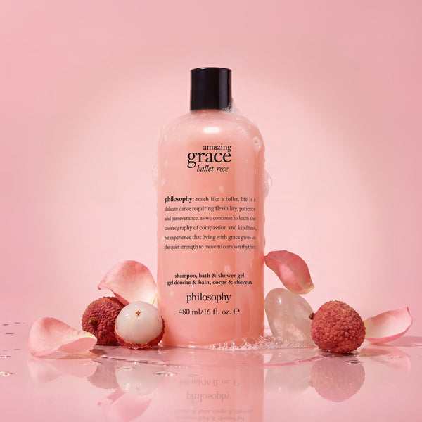 A bottle of Philosophy Amazing Grace Bullet Rose shampoo and bath & shower gel, styled with pink petals, lychee fruits, and ice on a reflective pink surface.